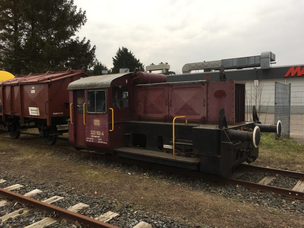 Die weinrote Köf mit braunen Güterwaggon. Dessen Dach ist schräg zugehend und kann geöffnet werden. Ein Deckel.