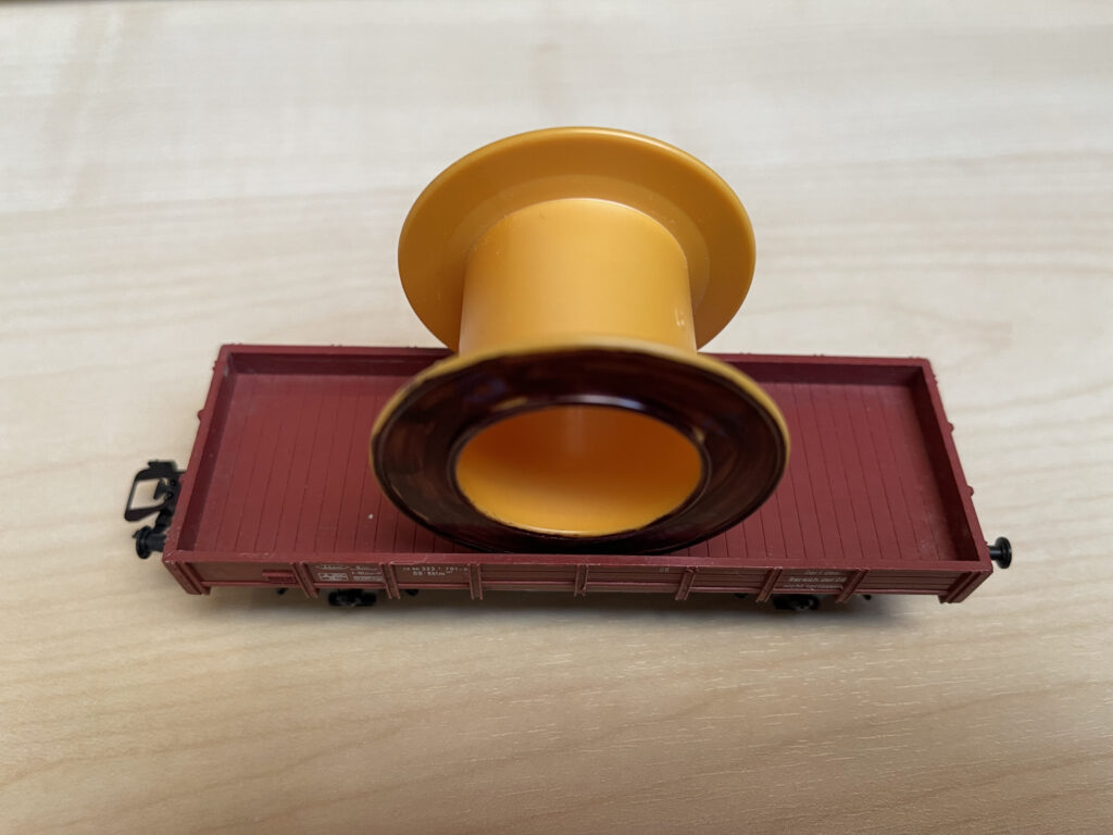 Auf einem kleinen braunen Niederbordwagen steht eine gelbe leere Rolle vom Klebeband. Die äußeren Seiten sind schwarz angemalt.