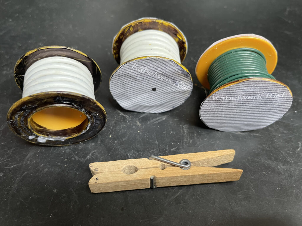 Auf einer schwarzen Gummimatte stehen die drei ehemals gelben Rollen. Auf zweien ist dickes weißes Kabel und auf einer grünes dünneres Kabel. Davor liegt eine Wäscheklammer aus Holz.