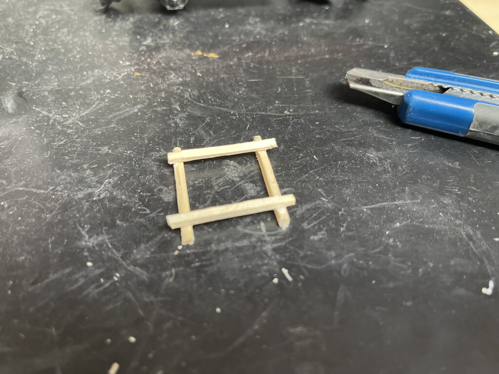 Auf einer schwarzen dreckigen Gummimatte liegen ein Schneidemesser, Cutter, und das Holzgestell aus Streichhölzern. Diese sind zu einem Viereck übereinander gelegt und verklebt.
