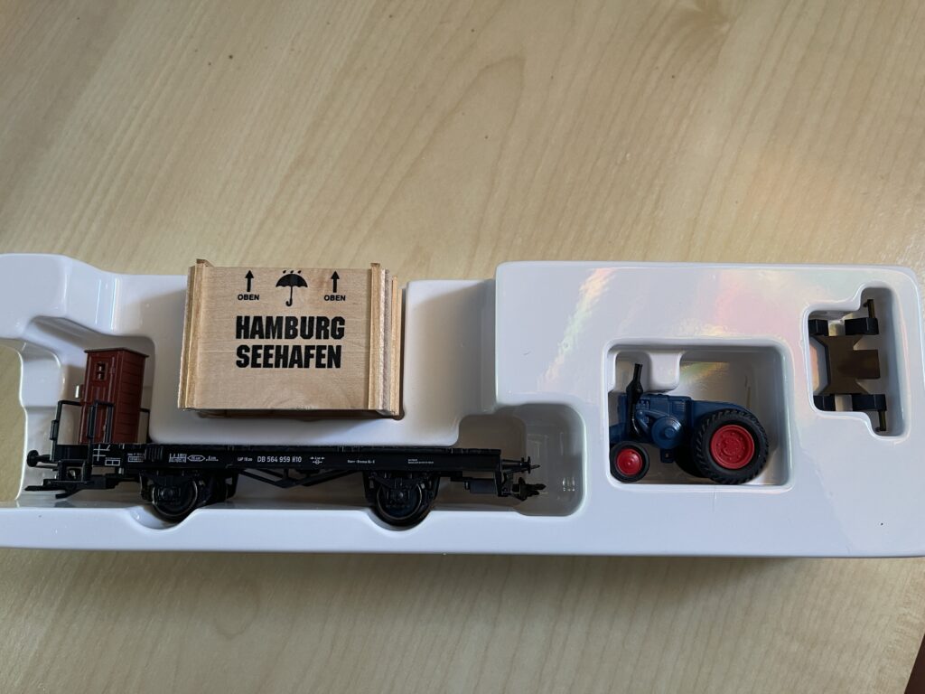 In der weißen Packung der Flachwagen, die hellbraune Holzkiste mit Aufschrift "Hamburg Seehafen" und Pfeilen für "Oben" und Regenschirm.
Dann der blaue Lanz Bulldog Traktor mit roten Felgen.
