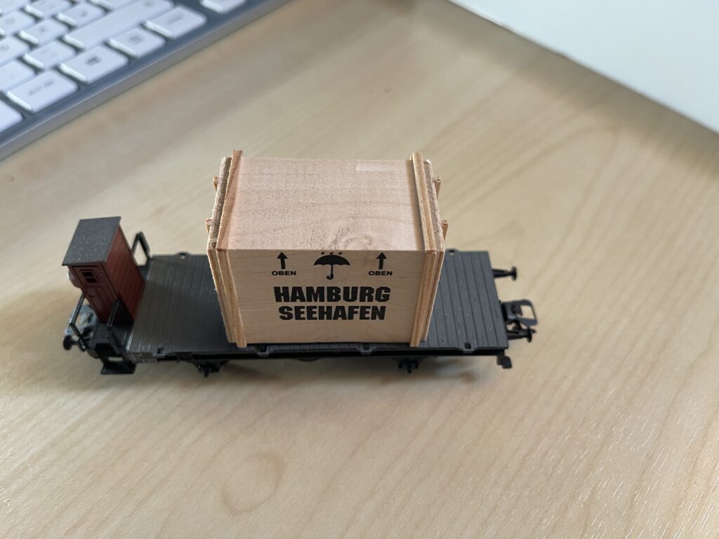 Der schwarze Flachwagen steht auf dem Tisch. Links das Bremserhäuschen aus Holz. Mitten auf dem Wagen ist eine hellbraune Holzkiste mit Aufschrift "Hamburg Seehafen" und Pfeilen für "Oben".