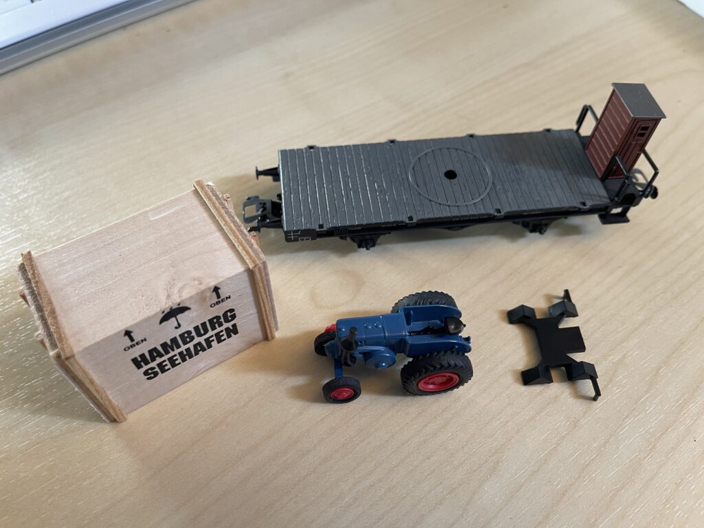 Zu sehen sind der schwarze Flachwagen, ein Eisenbahnwaggon ohne Seiten oder Rungen mit braunem Bremserhäuschen. Daneben steht eine hellbraune Holzkiste mit Aufschrift "Hamburg Seehafen" und Pfeilen für Oben und einem Regenschirm.
Vorn steht der blaue Traktor Lanz Bulldog mit schwarzen Reifen und roten Felgen. Daneben ein kleines schwarze X-förmiges Teil um den Traktor auf dem Wagen zu halten.