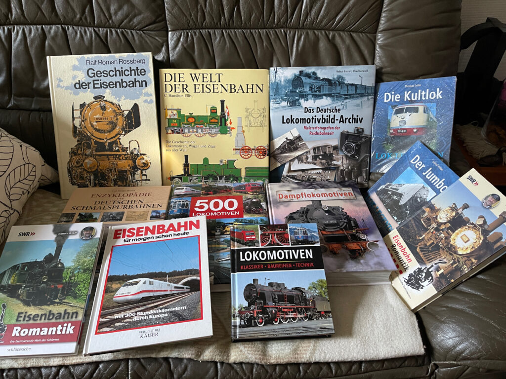 Auf einer dunklen Ledercouch liegen neben und übereinander 12 Eisenbahnbücher. Alle Cover zeigen Loks. Viele Dampfloks.