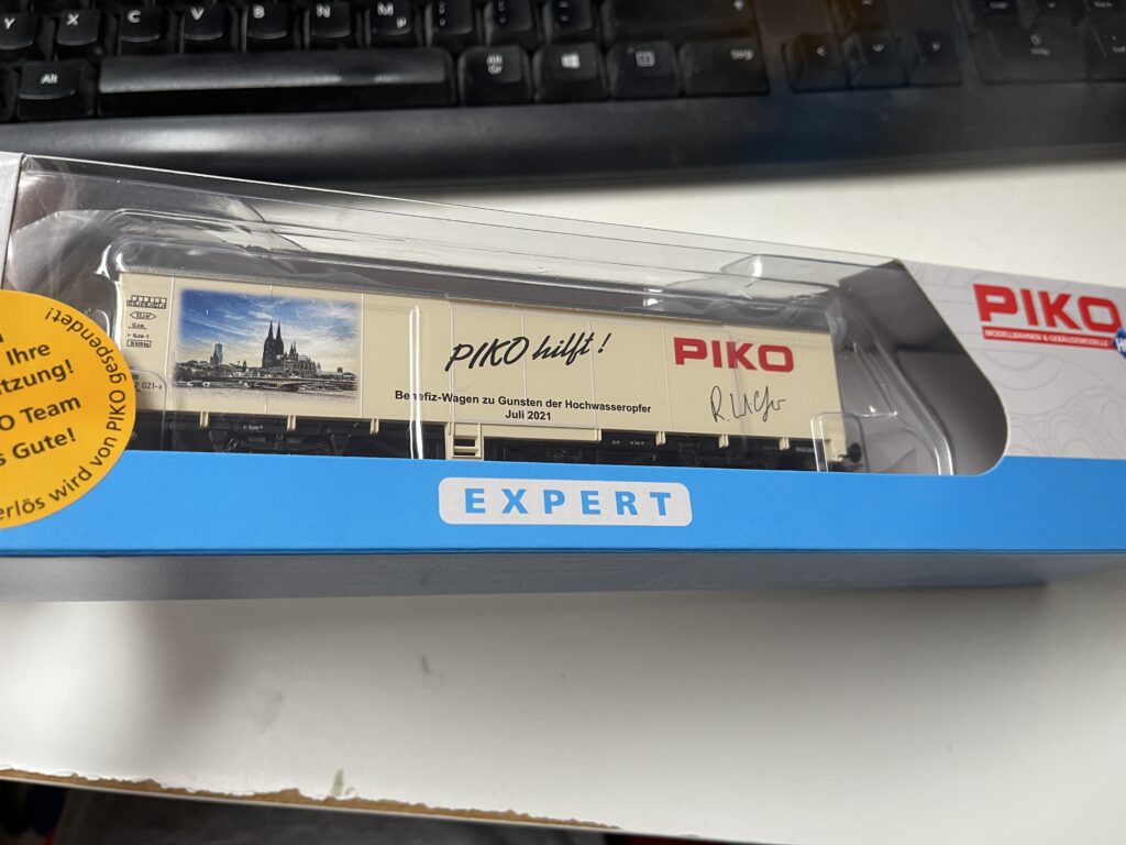 Der Benefiz-Wagen von PIKO kommt in einer weißen Packung mit blauem Rand unten. Der Wagen selbst ist leicht beige. Rechts in Rot das PIKO Logo und die Unterschrift des Geschäftsführers Wilfer.
Mittig PIKO hilft und da drunter der Text:
Benefiz-Wagen zu Gunsten der Hochwasseropfer Juli 2021.
Links ein Bild der Stadt Köln mit dem Dom.