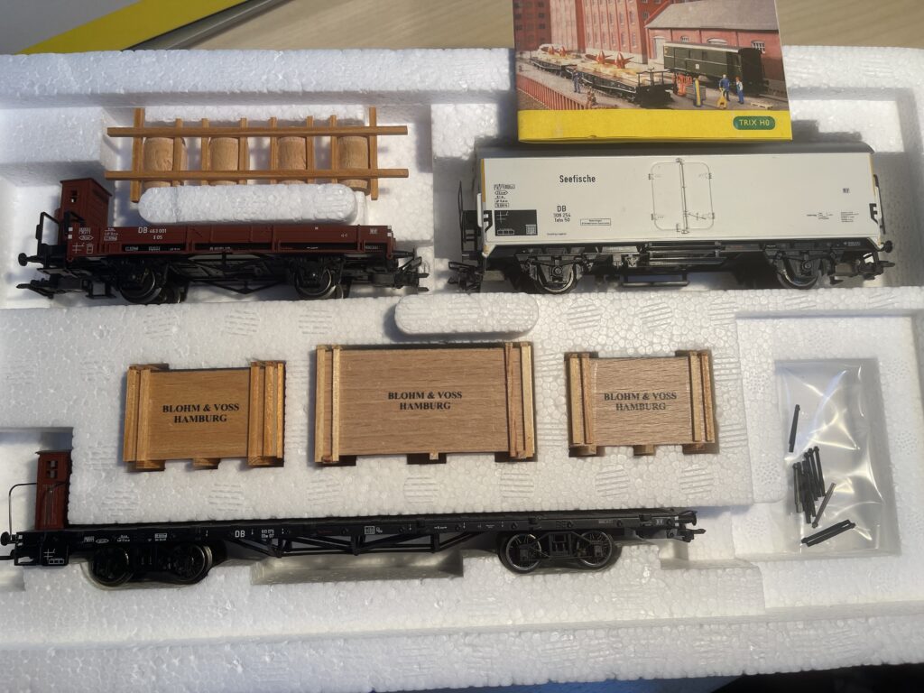 Die offene Packung. Oben ein Niederbordwagen. Darüber eine Halterung mit vier Fässern. Daneben ein weißer Kühlwagen Seefische.
Darunter drei große Holzkosten mit Aufschrift Blohm und Voss Hamburg.
Darunter ein flacher Wagen.