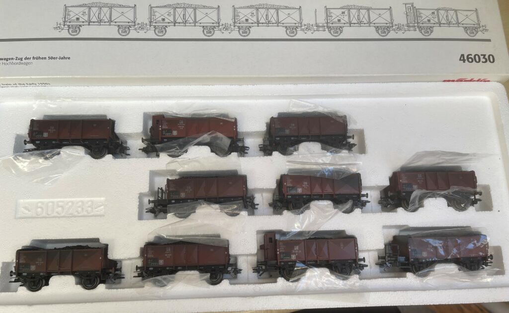 Oben ist gerade noch die Packung zu sehen mit fünf gezeichneten offenen Güterwagen. Darunter die offene Packung mit den Güterwagen. Deutlich geschwärzt. Die Wagen sind jeweils noch in Plastikfolie eingeschlagen.