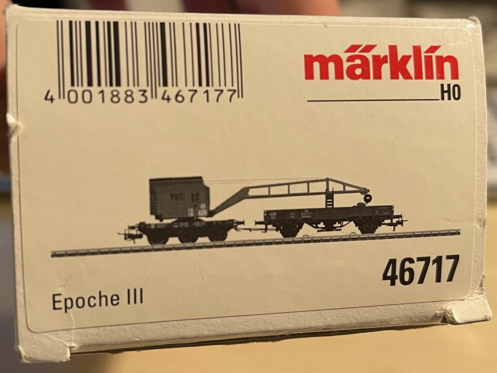 Weiße Packung mit rotem Märklin Logo.
In schwarz weiß der Kranwagen mit Niederbordwagen für den Kranausleger.
Darunter Epoche III und die Artikelnummer 46717.